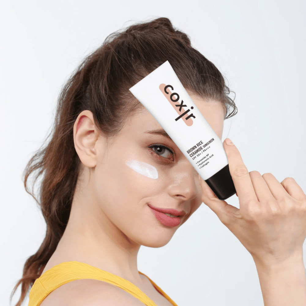 Femme avec crème solaire appliquée sur la joue tenant un tube Coxir Brown Rice Ceramide Sunscreen, SPF 50+ PA+++, vers le front et souriant légèrement.