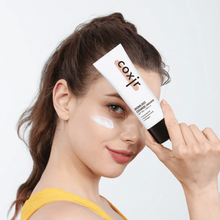 Femme avec crème solaire appliquée sur la joue tenant un tube Coxir Brown Rice Ceramide Sunscreen, SPF 50+ PA+++, vers le front et souriant légèrement.