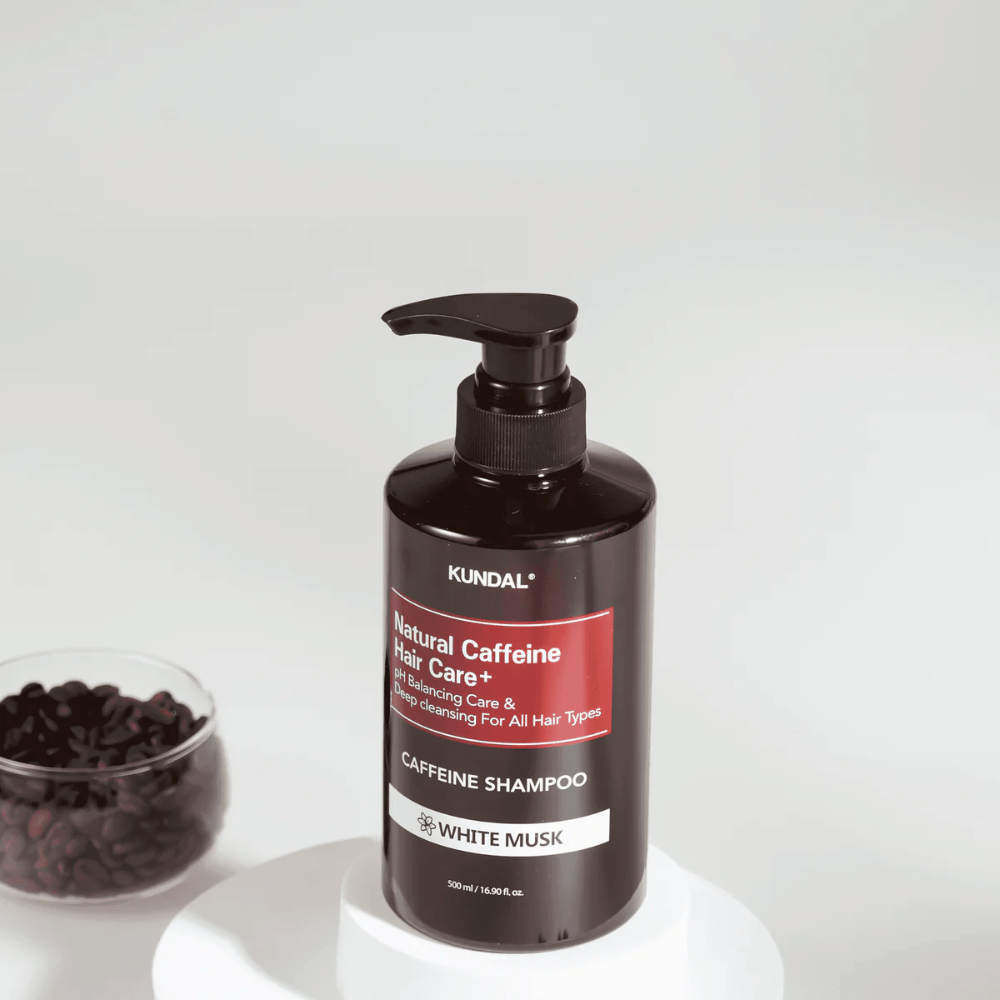 KUNDAL Natural Caffeine Hair Care+ Shampoo White Musk dans une bouteille avec pompe noire, 500 ml, placée à côté d'un bol de grains de café.