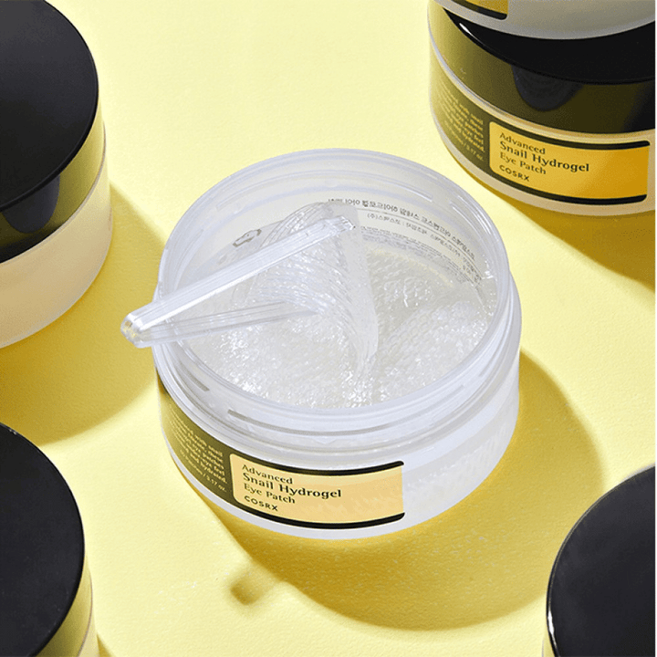 Un pot ouvert COSRX Advanced Snail Hydrogel Eye Patch avec des patchs pour les yeux transparents et une petite spatule, entouré de plusieurs pots fermés sur fond jaune.