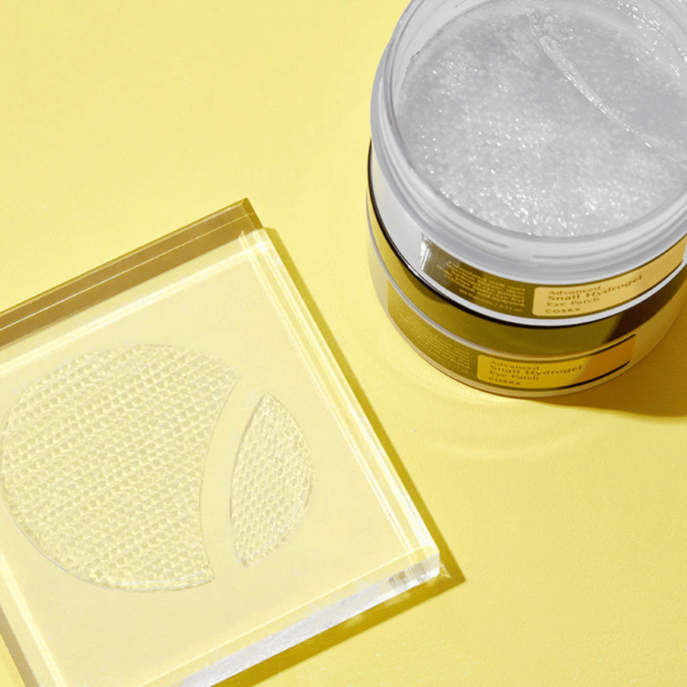 Un pot ouvert COSRX Advanced Snail Hydrogel Eye Patch se tient sur un fond jaune à côté d'un sous-verre transparent où deux patchs pour les yeux sont placés.