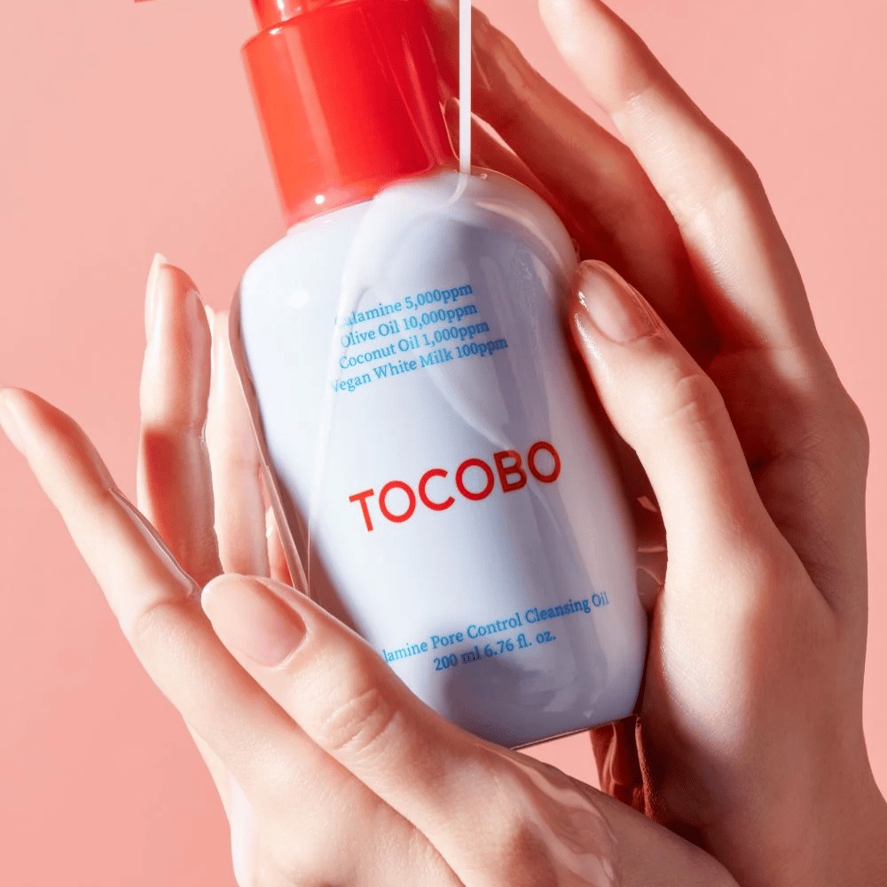 TOCOBO Calamine Pore Control Cleansing Oil tenus dans les mains contre un fond rose, la pompe rouge du flacon et la liste des ingrédients sont clairement visibles.