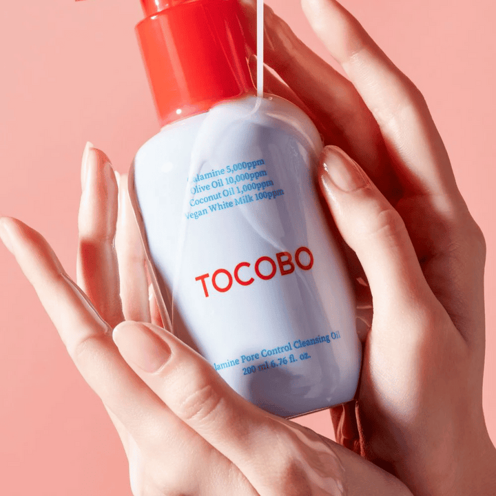 TOCOBO Calamine Pore Control Cleansing Oil tenus dans les mains contre un fond rose, la pompe rouge du flacon et la liste des ingrédients sont clairement visibles.