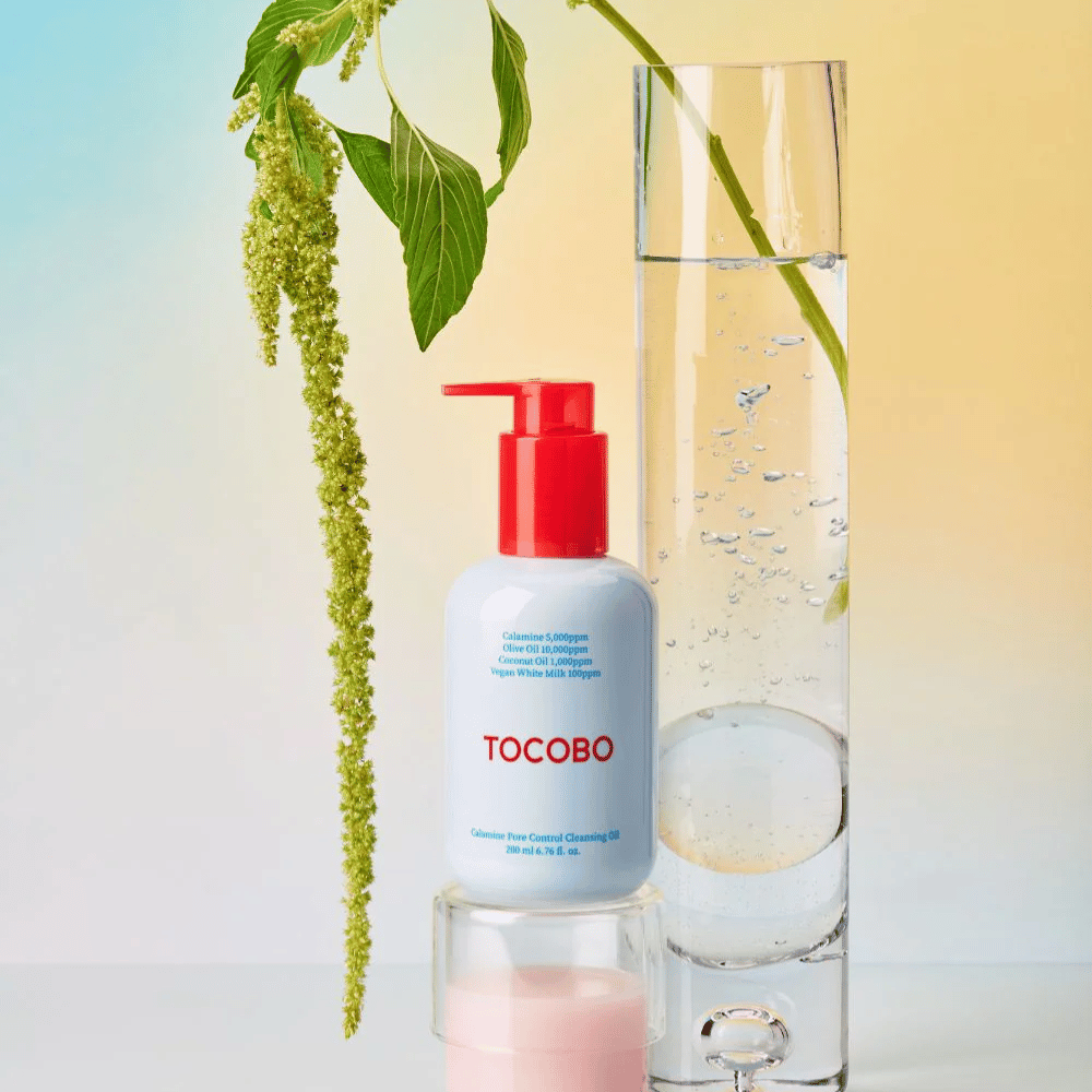 TOCOBO Calamine Pore Control Cleansing Oil avec pompe rouge se tient à côté d'un grand tube en verre rempli d'eau et d'une plante verte, sur un fond pastel clair.