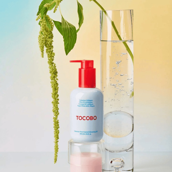 TOCOBO Calamine Pore Control Cleansing Oil avec pompe rouge se tient à côté d'un grand tube en verre rempli d'eau et d'une plante verte, sur un fond pastel clair.