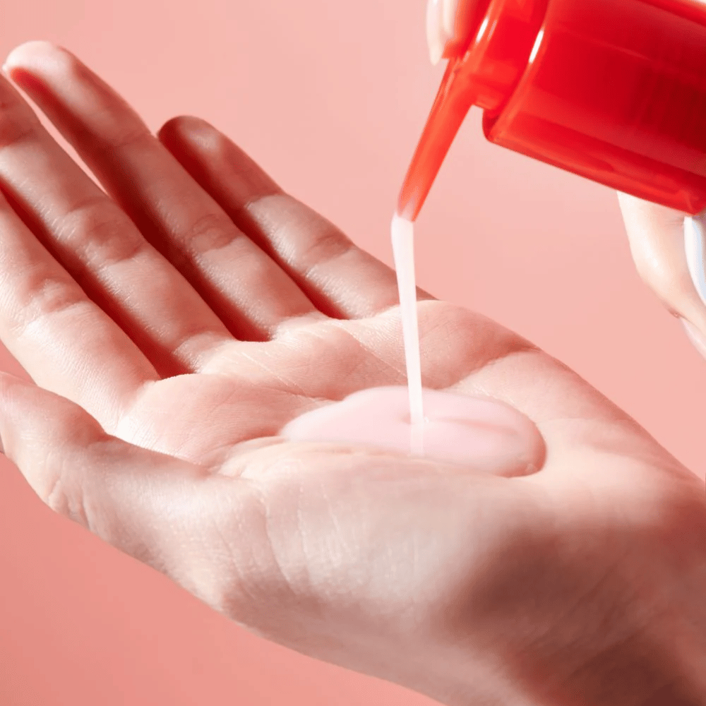 Une huile nettoyante rose pâle est appliquée d'un flacon pompe rouge dans une main ouverte contre un fond rose.