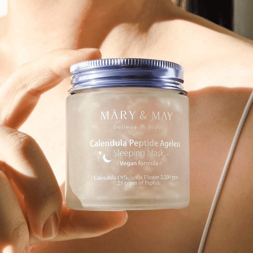 Une femme tient un pot de MARY & MAY Calendula Peptide Ageless Sleeping Mask. Le pot est transparent avec un couvercle violet et le texte "Calendula Peptide Ageless Sleeping Mask" est clairement visible. Le produit est un masque de nuit végan avec du calendula et des peptides, destiné à donner à la peau un éclat jeune pendant la nuit.