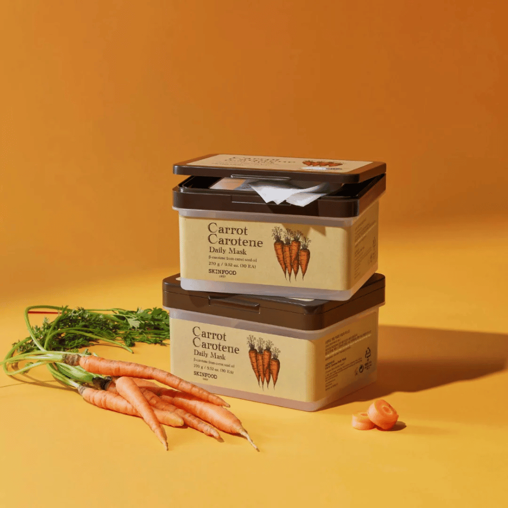 L'image montre trois emballages empilés de "Carrot Carotene Daily Mask" de SKINFOOD, placés sur un fond orange avec des carottes fraîches. Chaque boîte contient des masques faciaux enrichis en β-carotène provenant de l'huile de graines de carotte, conçus pour un usage quotidien pour nourrir et soigner la peau.
