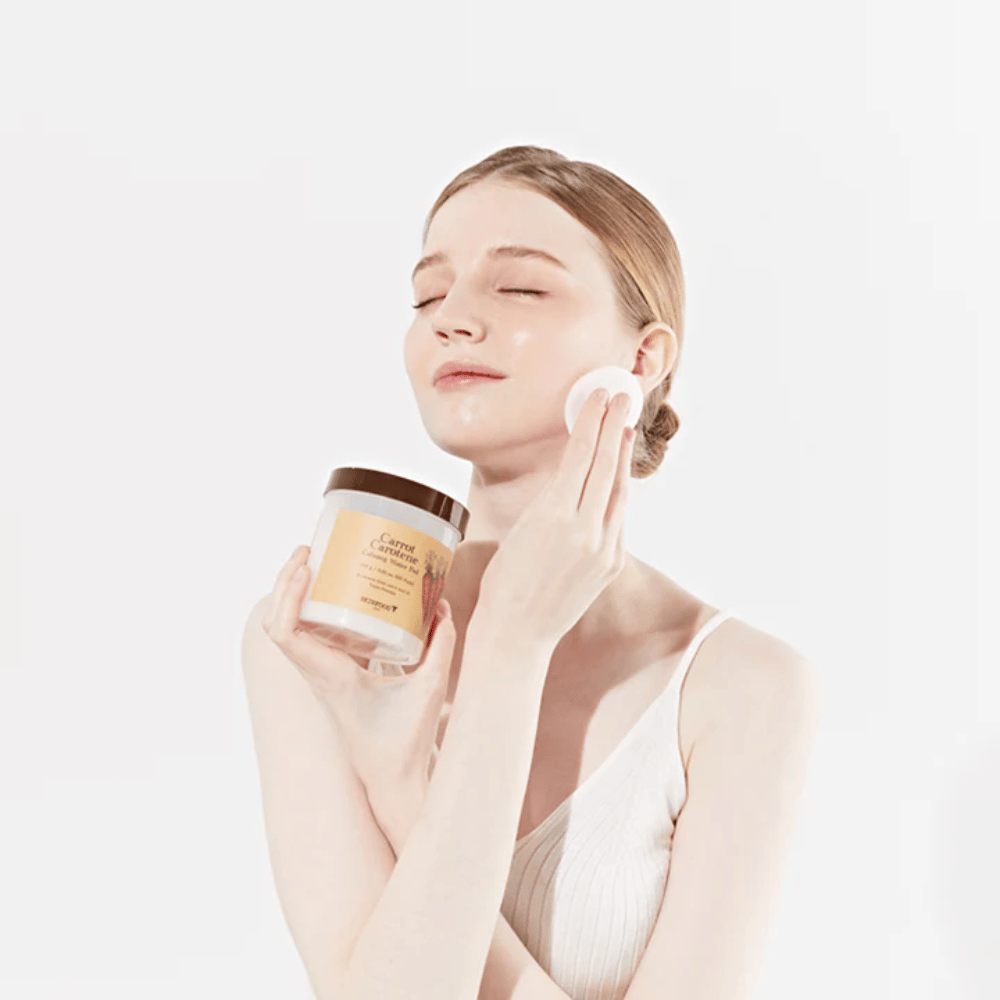 Une femme aux cheveux attachés applique un produit de soin de la peau sur son visage avec un tampon en coton. Elle tient un grand pot marqué "Carrot Carotene Calming Water Pad". Ses yeux fermés et son expression paisible transmettent un sentiment de détente et de plaisir dans la routine de soins de la peau.