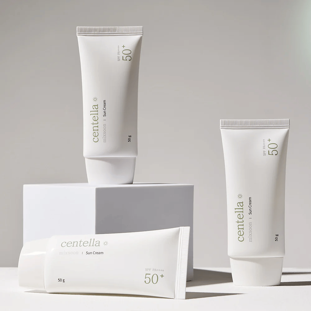 Trois emballages de Centella Sun Cream SPF 50+ PA++++ disposés esthétiquement sur des podiums blancs. Deux tubes sont debout et un est couché, tous avec un design blanc minimaliste et des détails de texte verts.
