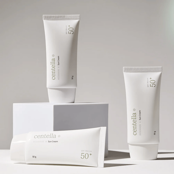 Trois emballages de Centella Sun Cream SPF 50+ PA++++ disposés esthétiquement sur des podiums blancs. Deux tubes sont debout et un est couché, tous avec un design blanc minimaliste et des détails de texte verts.