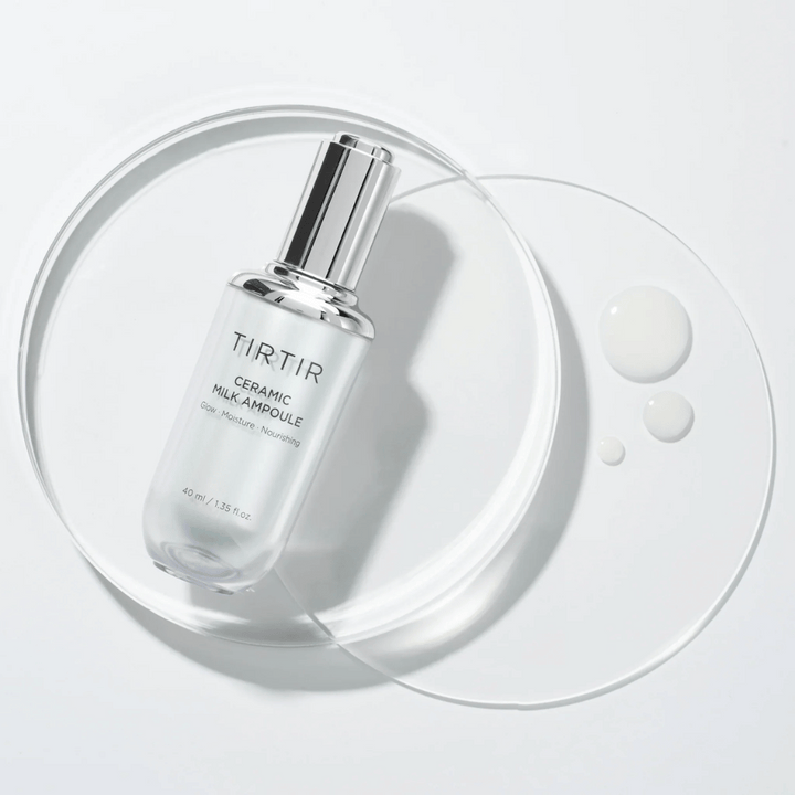 Une bouteille transparente TIRTIR Ceramic Milk Ampoule sur une plaque de verre avec des gouttes, contre un fond blanc, met en valeur l'éclat, l'hydratation et la nutrition.