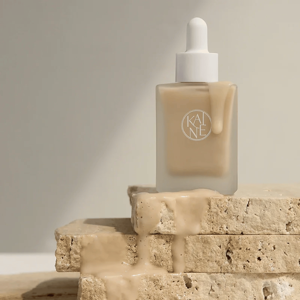 KANE Chaga Collagen Charging Serum dans une bouteille en verre dépoli avec pipette. Texture crémeuse coulant sur des pierres naturelles dans une lumière douce et minimaliste.