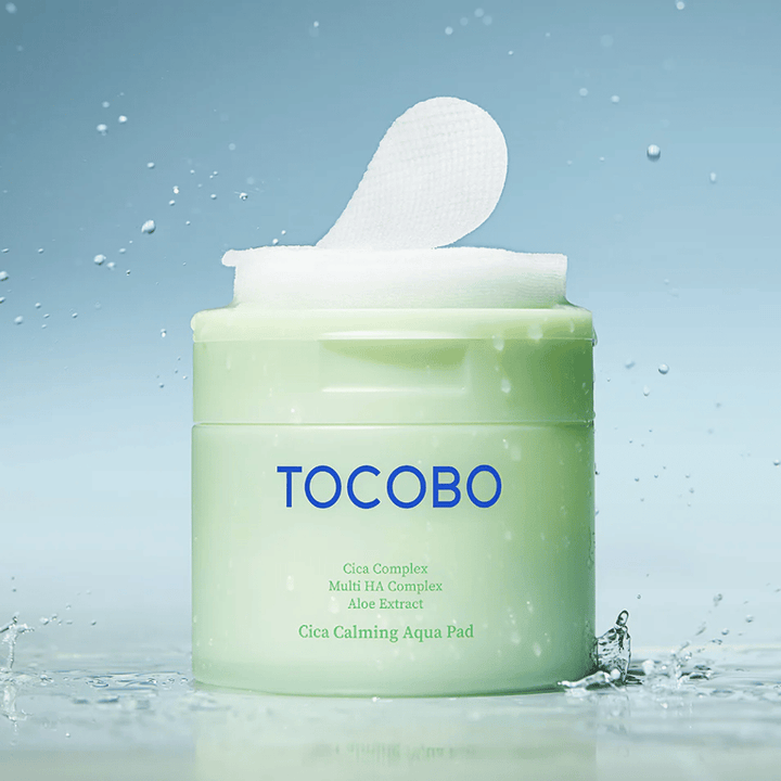 Pot ouvert de TOCOBO Cica Calming Aqua Pad d’où un pad toner visage dépasse de l’emballage. Des gouttes d’eau autour du produit renforcent la sensation d’hydratation intense et de fraîcheur.