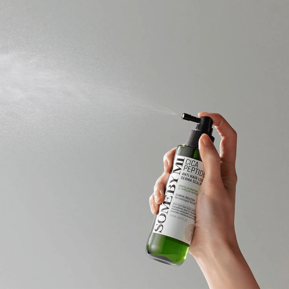 Main pulvérisant SOME BY MI Cica Peptide Anti Hair Loss Derma Scalp Tonic d'une bouteille verte avec un embout noir et une étiquette blanche sur fond gris.