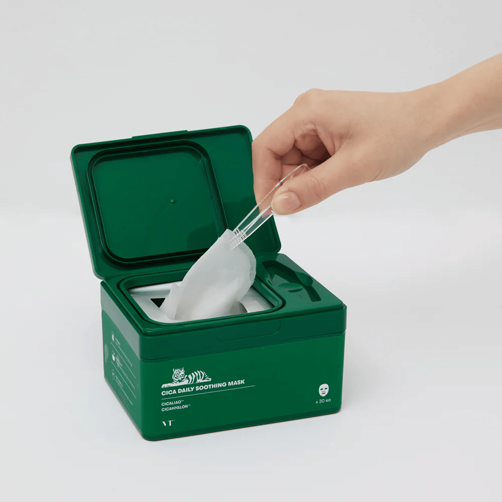 Une main utilise une pince pour retirer un Masque tissu d'un récipient vert marqué "Cica Daily Soothing Mask".