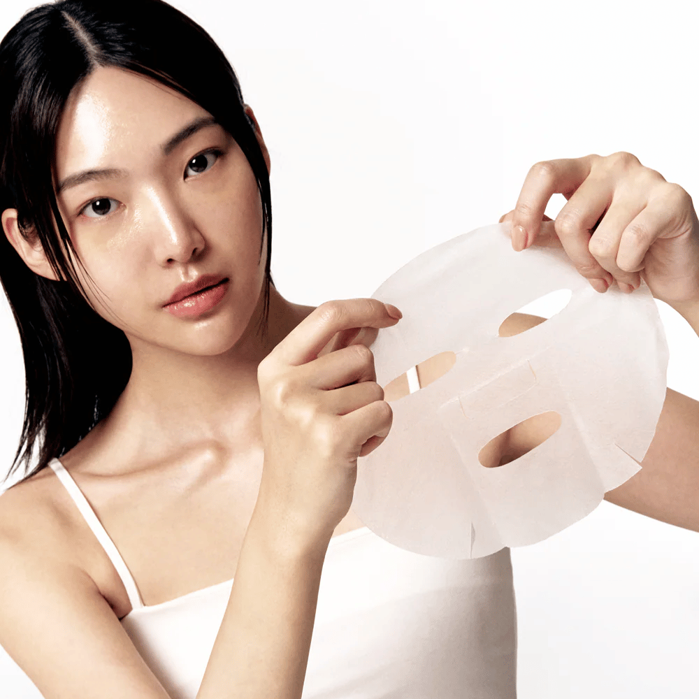 Femme avec une peau propre et hydratée tient un Masque tissu blanc devant elle, prête à l'appliquer sur le visage.