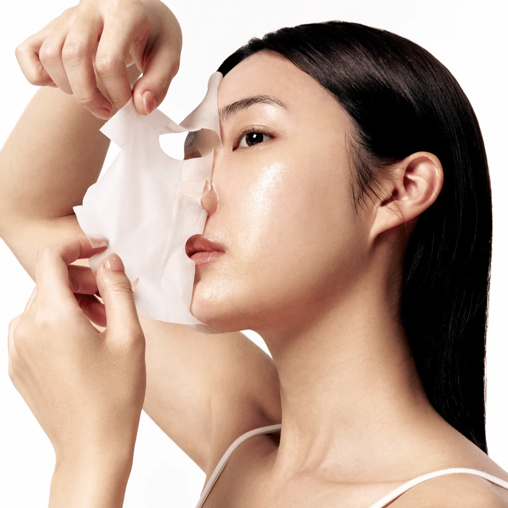 Femme avec une peau lisse et hydratée retire un Masque tissu blanc de son visage, révélant un teint éclatant et uniforme.