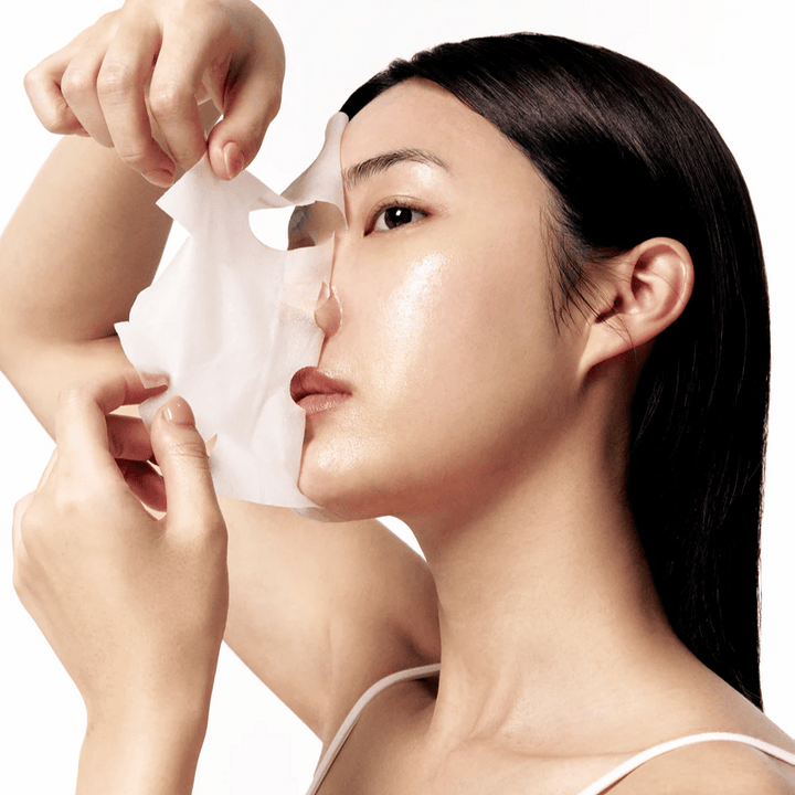 Femme avec une peau lisse et hydratée retire un Masque tissu blanc de son visage, révélant un teint éclatant et uniforme.