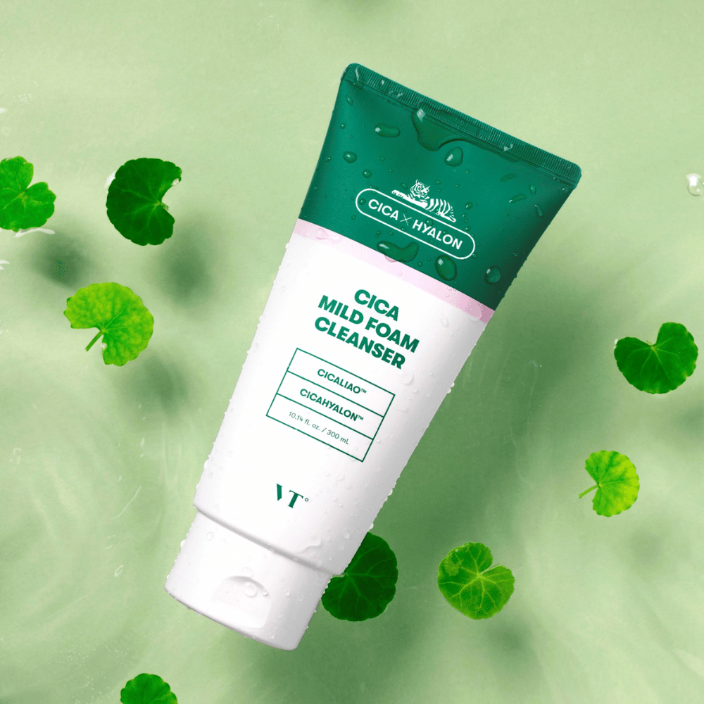 VT Cica Mild Foam Cleanser couché sur un fond vert clair avec des gouttes d'eau sur l'emballage et entouré de feuilles vertes de centella asiatica, mettant en valeur les propriétés apaisantes du produit.
