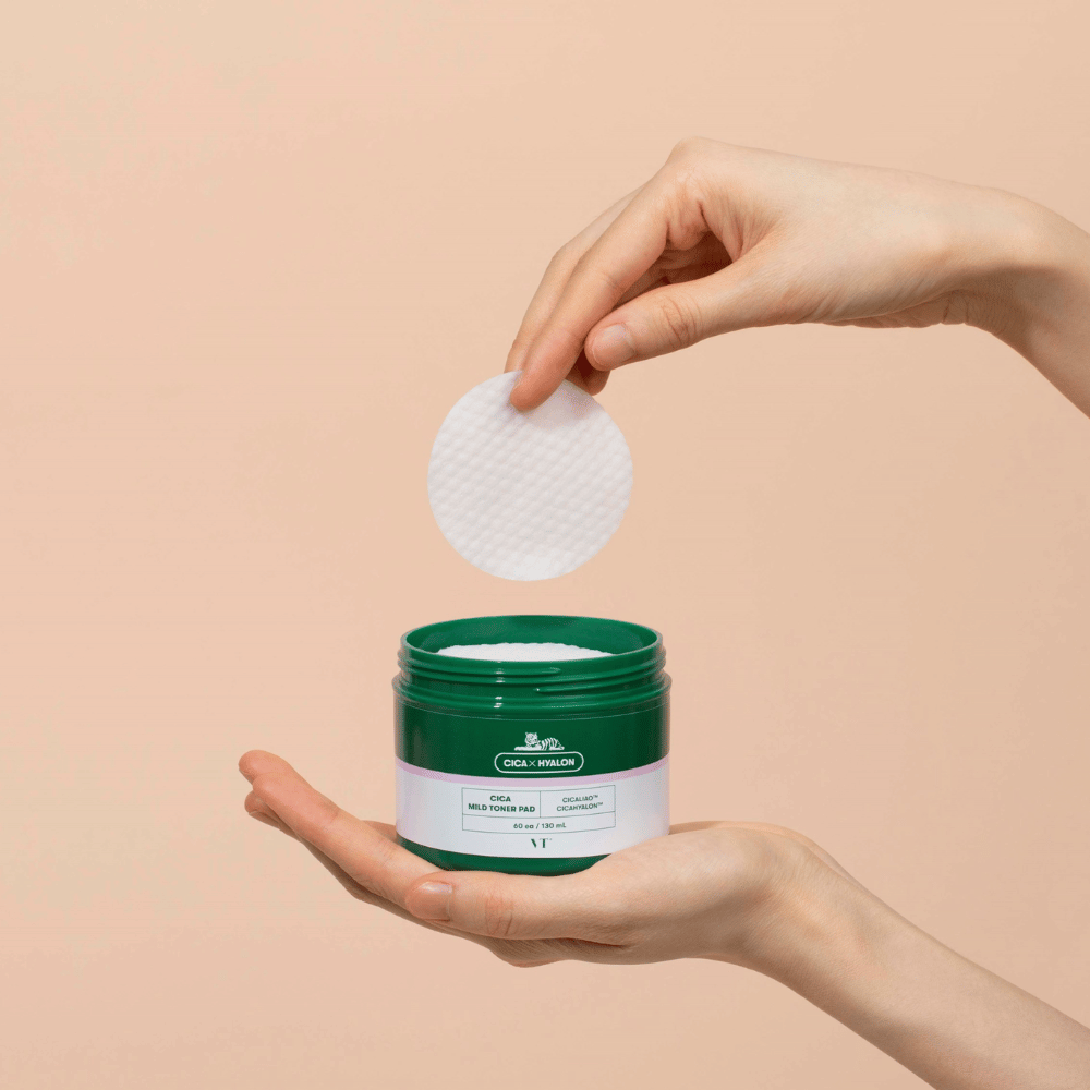 Main tenant un pot vert avec VT Cica Mild Toner Pad et soulevant un tampon de toner rond et blanc au-dessus du pot sur fond beige.