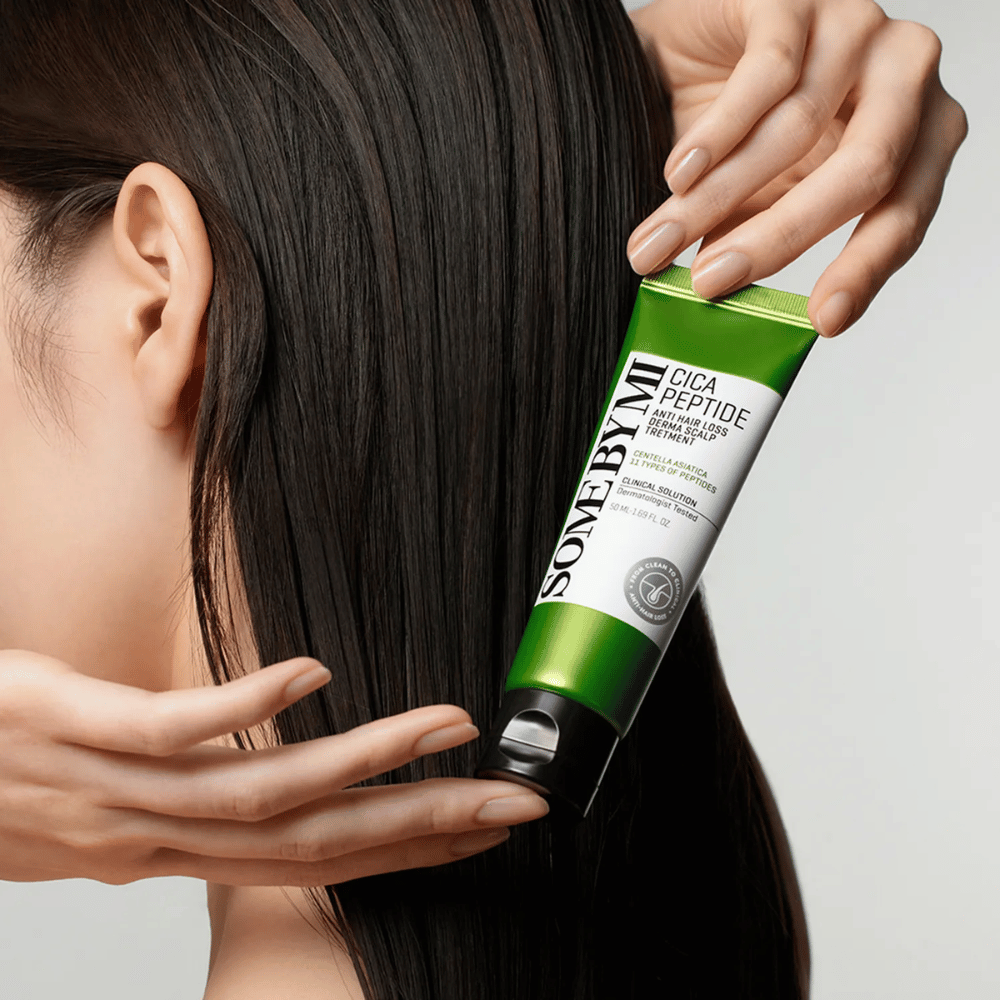 Une femme applique SOME BY MI Cica Peptide Anti Hair Loss Derma Scalp Treatment dans de longs cheveux foncés avec un tube vert et blanc.