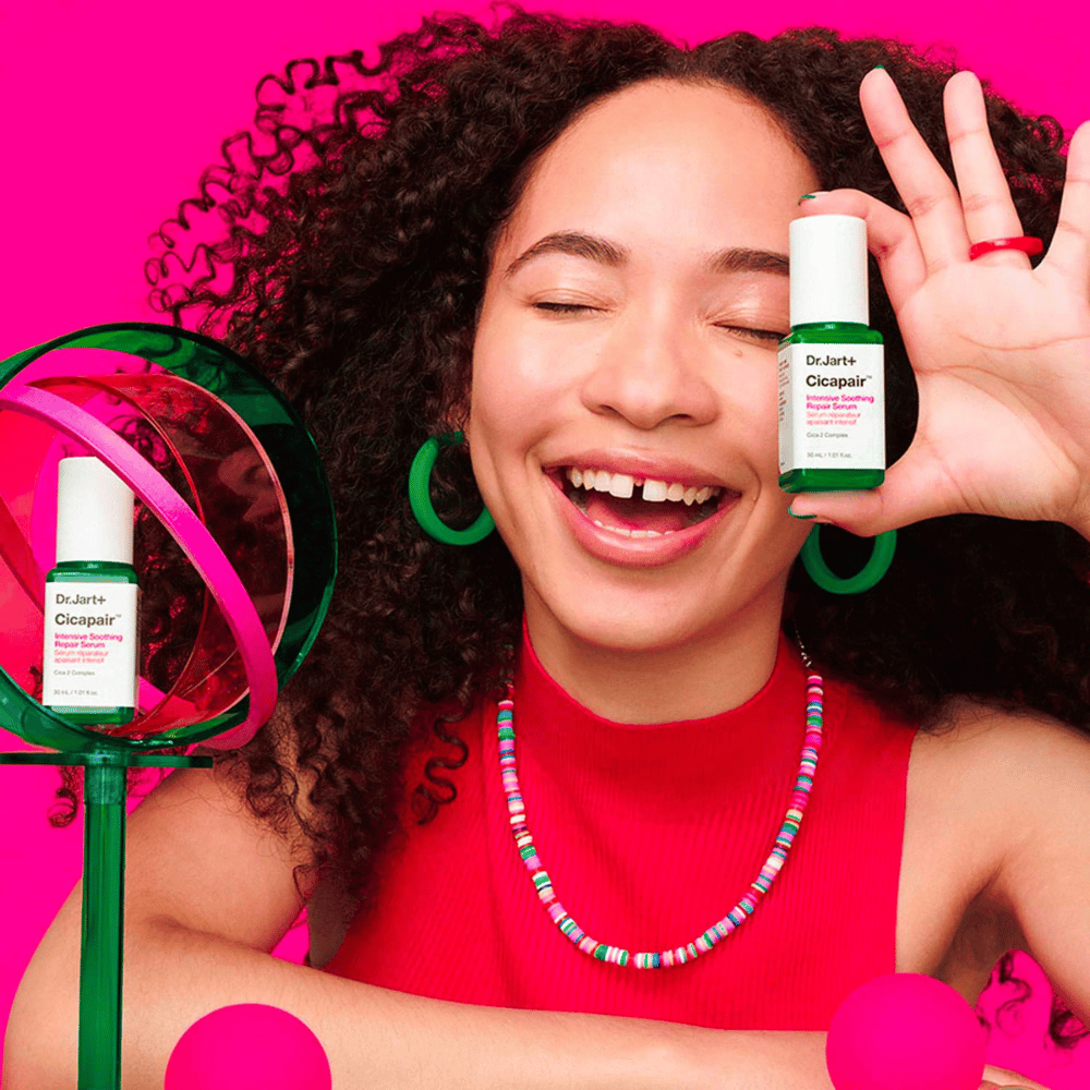 Une femme sourit largement tout en tenant Dr.Jart+ Cicapair Intensive Soothing Repair Serum devant son visage. Elle porte des boucles d'oreilles vertes et un collier rouge. L'arrière-plan est rose clair et décoré de cercles verts et roses.