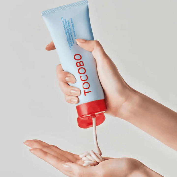 Une personne tient un tube de TOCOBO Coconut Clay Cleansing Foam. Le tube est bleu clair avec un bouchon rouge et le texte "TOCOBO" sur le devant. Le produit est pressé hors du tube et prend la forme d'un shampooing dans la main. L'arrière-plan est neutre et met l'accent sur le produit.
