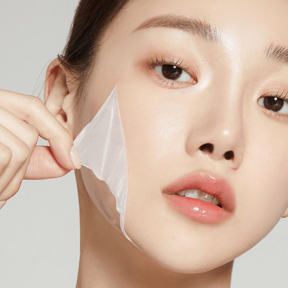 L'image montre une jeune femme en train de retirer un masque facial fin, mettant en valeur sa peau hydratée et douce. Le masque épouse les contours du visage, donnant l'impression d'un produit de soin de la peau efficace et doux. Son expression est détendue.