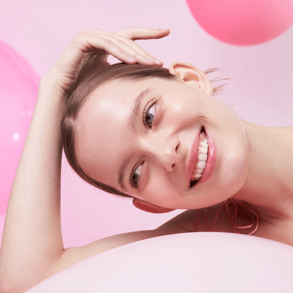 Une femme sourit et repose sa tête sur sa main, entourée de ballons roses. Sa peau semble éclatante et naturellement saine.