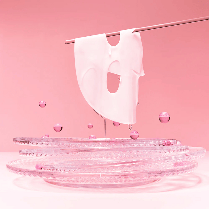 Un masque en hydrogel est suspendu à une tige, dégoulinant de sérum. Le fond est rose et le miroir en bas reflète les bulles roses qui entourent le masque.
