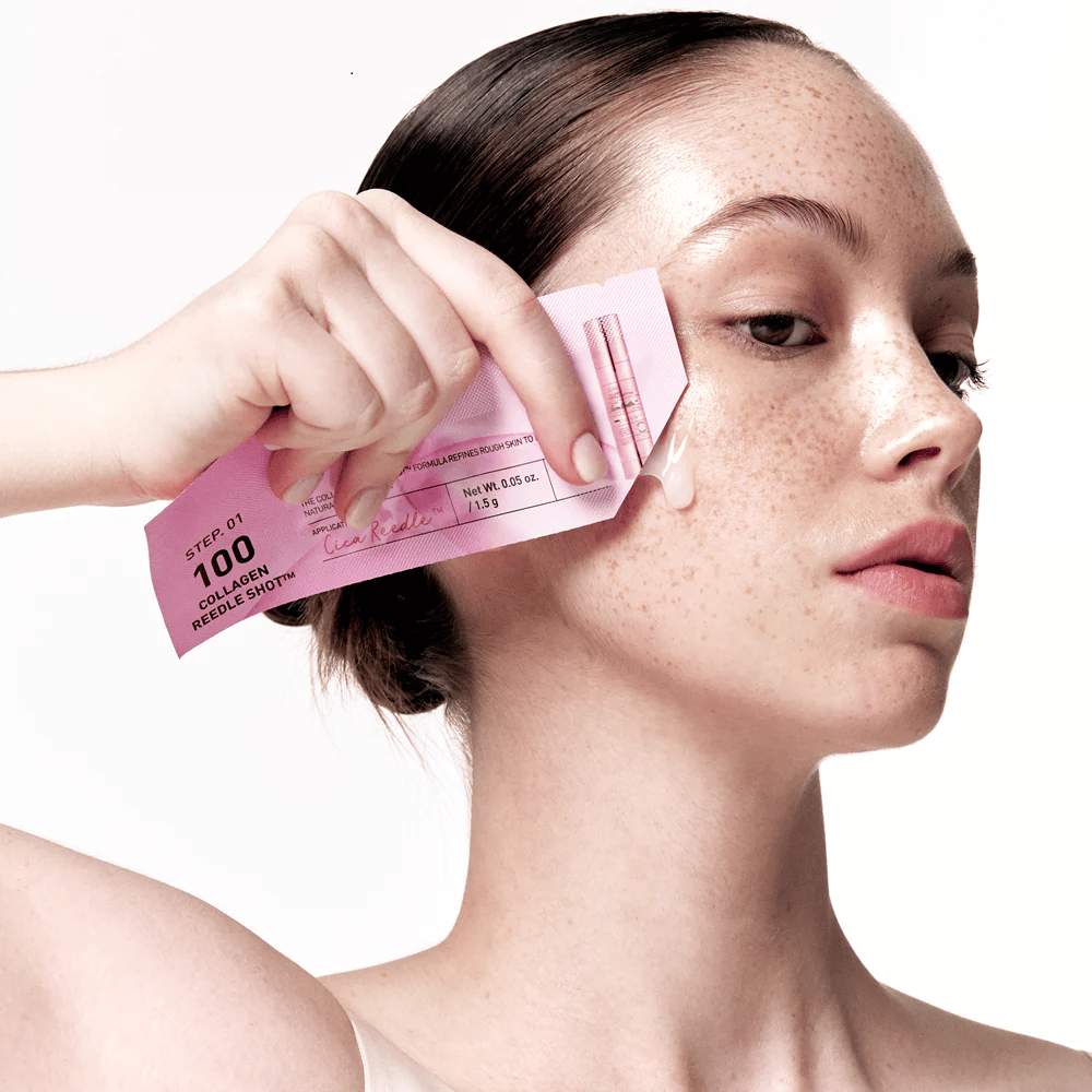 Une femme applique l'étape 1 de Collagen Reedle Shot 100 avec un emballage rose à usage unique sur la joue. Un gel clair est visible sur la peau, marquant la première étape du traitement de soin de la peau en deux étapes.
