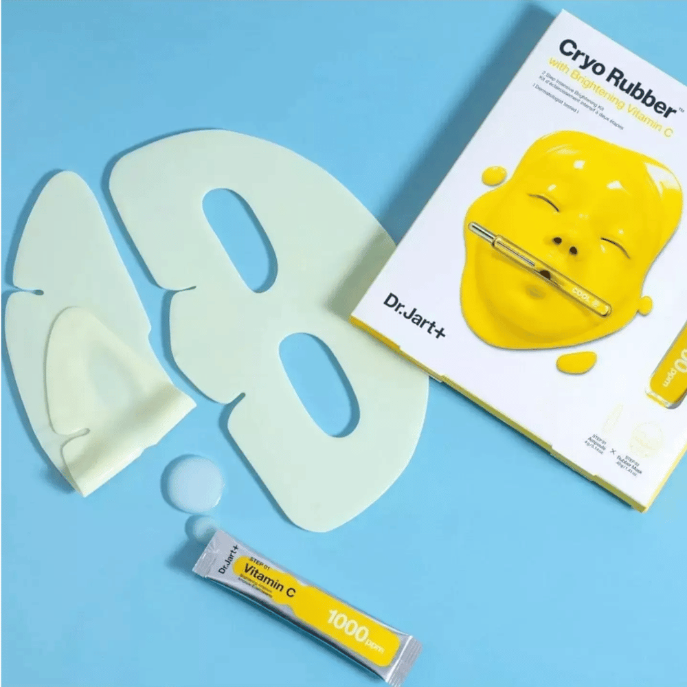 Image de Dr.Jart+'s produit de beauté 'Cryo Rubber™ avec Vitamine C Éclaircissante'. Sur un fond bleu, on voit un masque facial en caoutchouc blanc, un petit tube marqué 'Vitamine C 1000ppm', et une boîte avec le nom du produit et l'image du masque jaune.