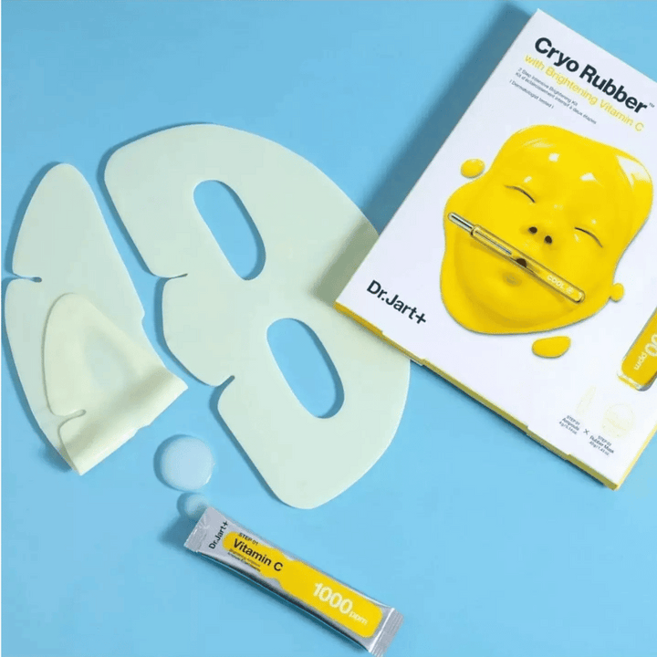 Image de Dr.Jart+'s produit de beauté 'Cryo Rubber™ avec Vitamine C Éclaircissante'. Sur un fond bleu, on voit un masque facial en caoutchouc blanc, un petit tube marqué 'Vitamine C 1000ppm', et une boîte avec le nom du produit et l'image du masque jaune.