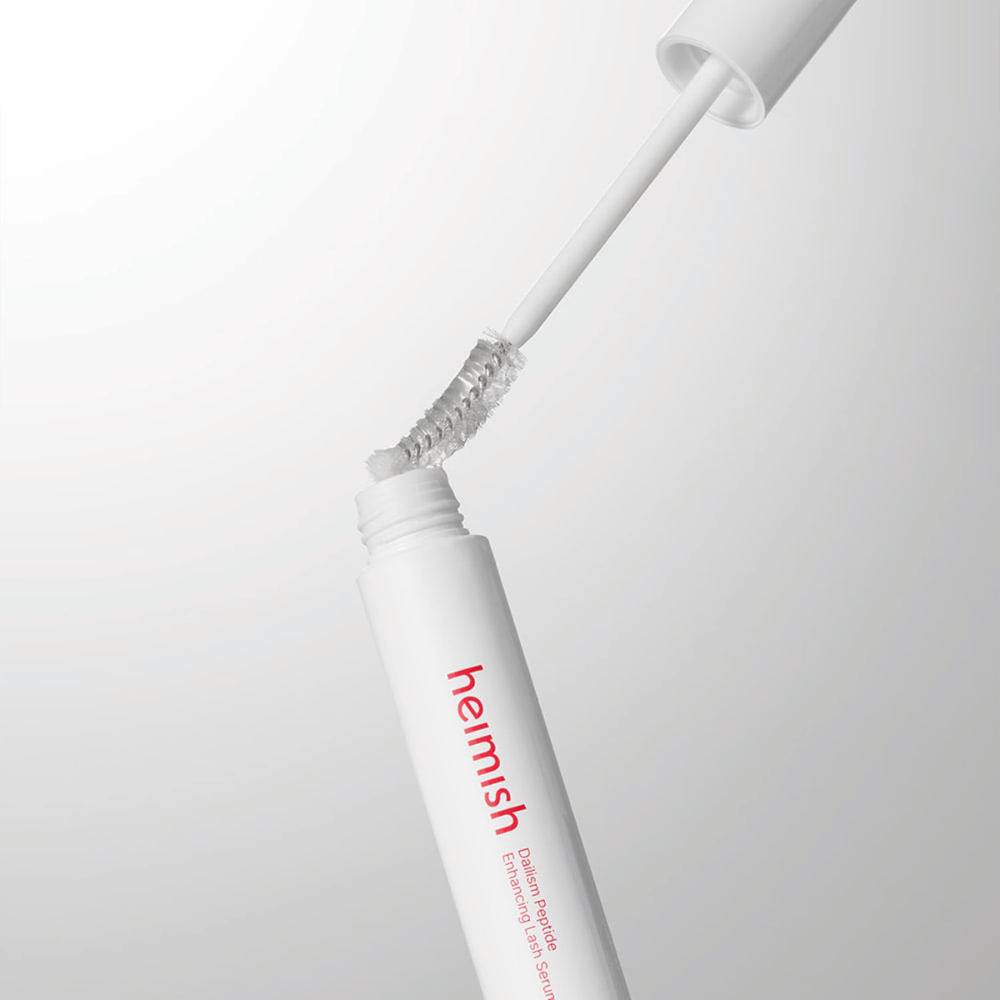Gros plan sur l’applicateur de Heimish Dailism Peptide Enhancing Lash Serum. La brosse est retirée du contenant blanc et montre la formule transparente du sérum qui s’applique sur les cils.