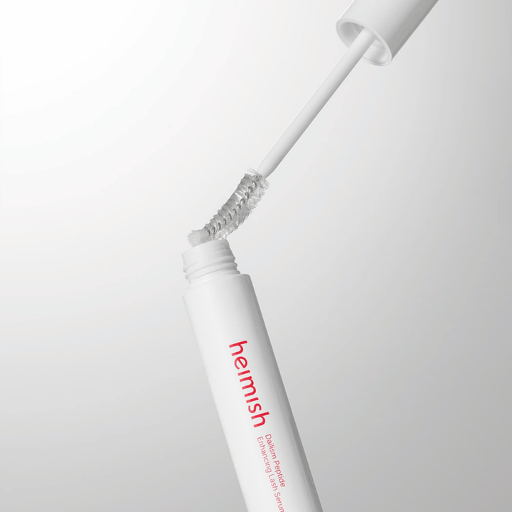 Gros plan sur l’applicateur de Heimish Dailism Peptide Enhancing Lash Serum. La brosse est retirée du contenant blanc et montre la formule transparente du sérum qui s’applique sur les cils.