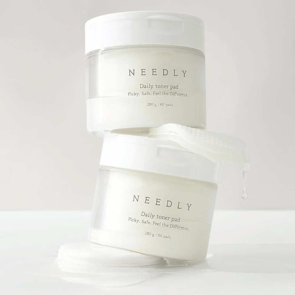 Deux pots avec Needly Daily Toner Pad empilés l'un sur l'autre. Les pots sont blancs avec un texte minimaliste et contiennent chacun 60 tampons de toner. Un tampon de toner est partiellement à l'extérieur du pot supérieur avec du liquide qui en goutte.