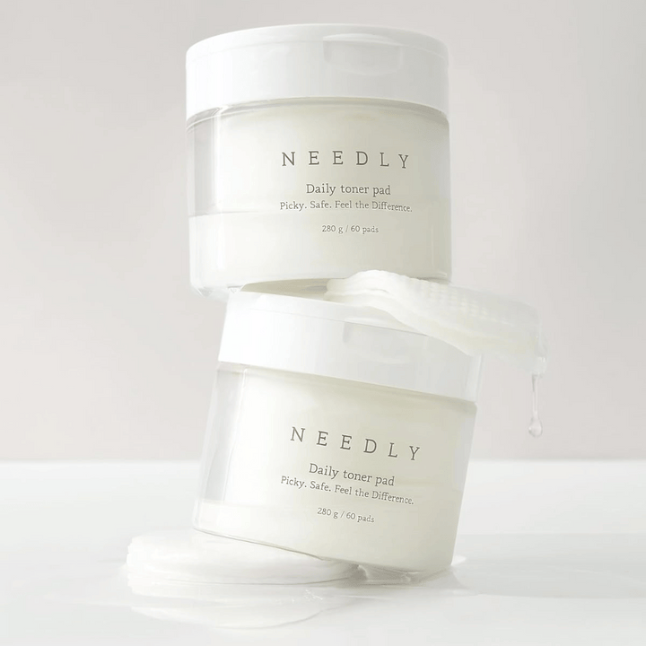 Deux pots avec Needly Daily Toner Pad empilés l'un sur l'autre. Les pots sont blancs avec un texte minimaliste et contiennent chacun 60 tampons de toner. Un tampon de toner est partiellement à l'extérieur du pot supérieur avec du liquide qui en goutte.