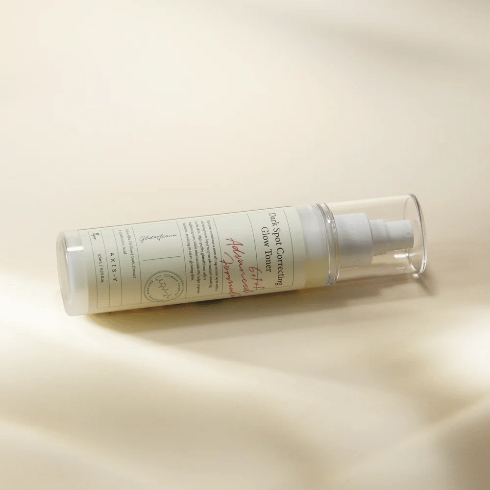 Une bouteille couchée de "AXIS-Y Dark Spot Correcting Glow Toner" placée sur une surface claire. La bouteille est transparente avec une étiquette blanche et a une pompe à spray blanche. L'étiquette montre des informations sur les propriétés éclaircissantes et hydratantes du produit et contient des ingrédients clés tels que le glutathion et la niacinamide.