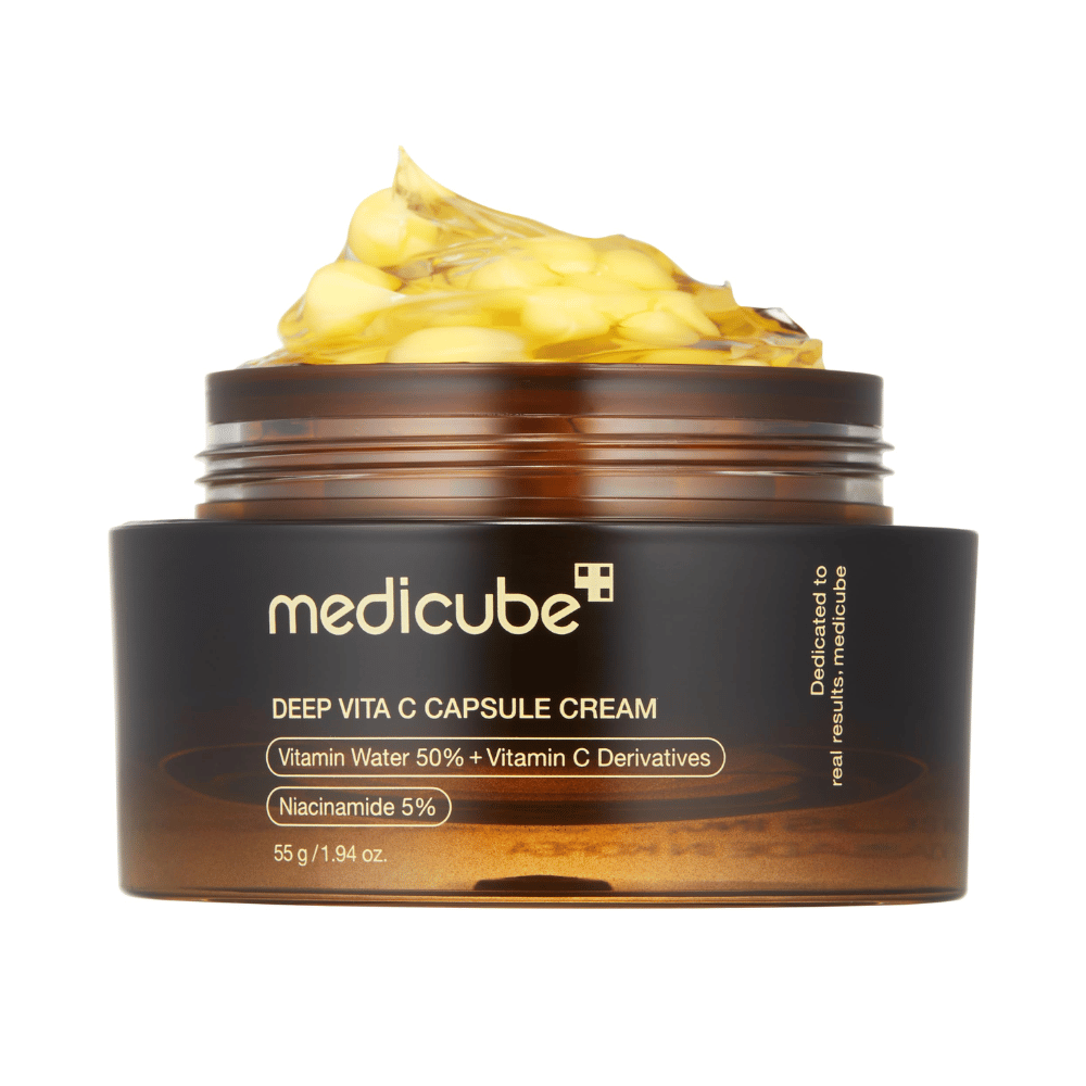 Un gros plan de "Medicube Deep Vita C Capsule Cream," montrant la consistance de la crème et les capsules de vitamine C. L'emballage est brun foncé avec du texte doré et décrit le contenu en eau vitaminée (50 %) et niacinamide (5 %), offrant une présentation visuelle des propriétés hydratantes et améliorant la peau du produit.