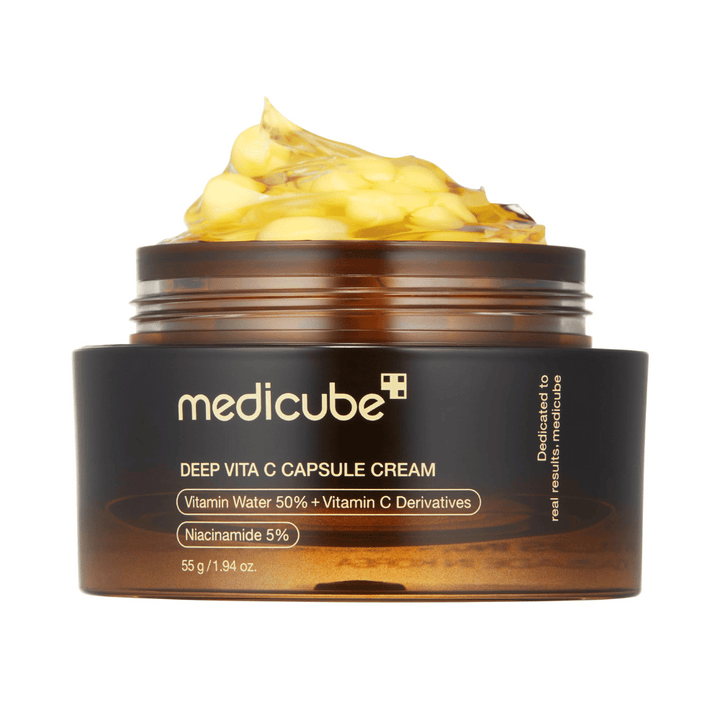 Un gros plan de "Medicube Deep Vita C Capsule Cream," montrant la consistance de la crème et les capsules de vitamine C. L'emballage est brun foncé avec du texte doré et décrit le contenu en eau vitaminée (50 %) et niacinamide (5 %), offrant une présentation visuelle des propriétés hydratantes et améliorant la peau du produit.