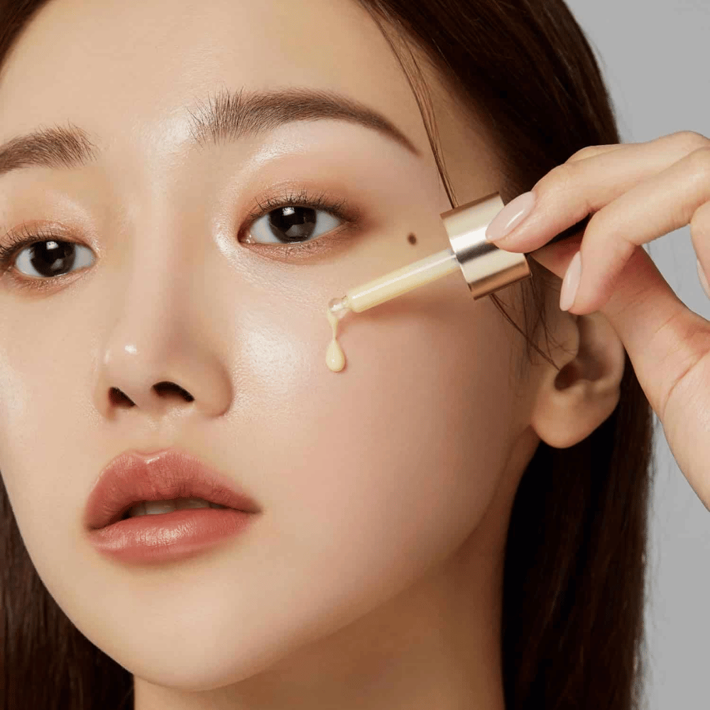 Gros plan d'une femme appliquant Medicube Deep Vita C Ampoule avec une pipette dorée. Une goutte de sérum est sur le point d'atteindre sa peau, qui a un éclat naturel. Parfait pour donner un teint plus lumineux et plus uniforme.