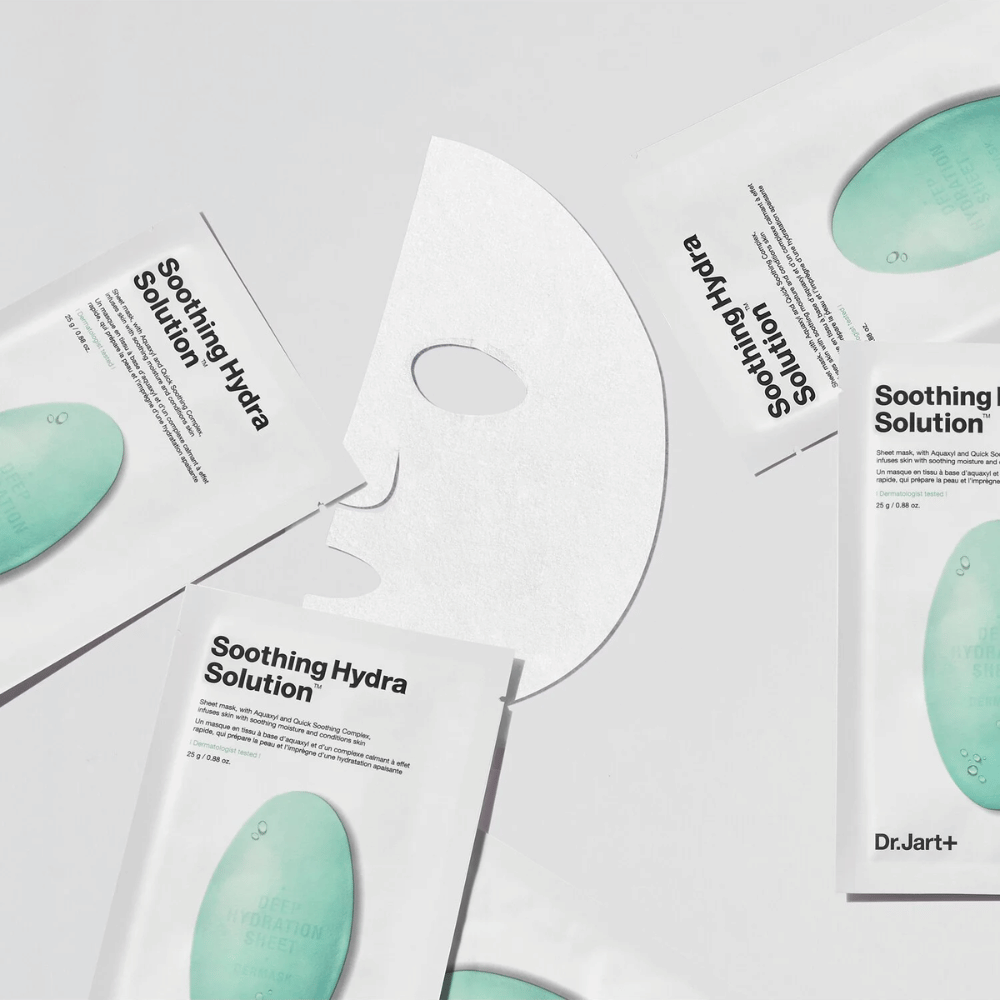 Plusieurs emballages de masques faciaux "Soothing Hydra Solution" de Dr.Jart+ sont éparpillés sur une surface claire. Un masque déplié est placé au centre, montrant sa structure blanche et perforée.