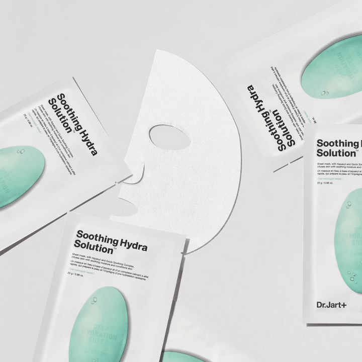 Plusieurs emballages de masques faciaux "Soothing Hydra Solution" de Dr.Jart+ sont éparpillés sur une surface claire. Un masque déplié est placé au centre, montrant sa structure blanche et perforée.