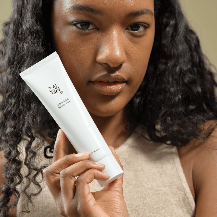 Une femme aux cheveux bouclés tient un tube "Beauty of Joseon Dynasty Cream" près de son visage, mettant en valeur le produit. La crème, présentée dans un tube blanc élégant, est conçue pour nourrir et rajeunir la peau, soulignant son attrait pour différents types de peau.