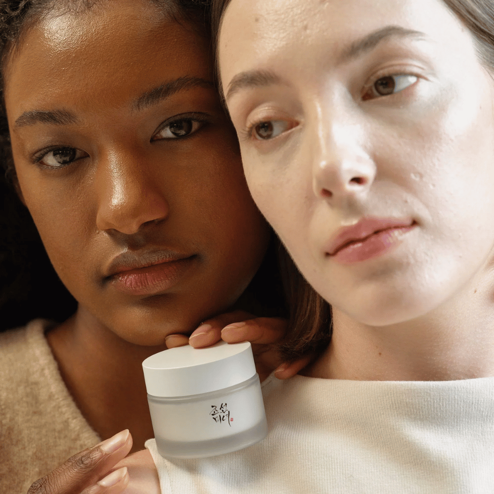 Deux femmes, l'une avec une peau plus foncée et l'autre avec une peau plus claire, se tiennent près l'une de l'autre avec un focus sur leurs visages et une crème pour la peau de "Beauty of Joseon". La crème est placée devant elles, soulignant son utilisation pour différents types de peau et sa capacité à apporter éclat et nutrition.