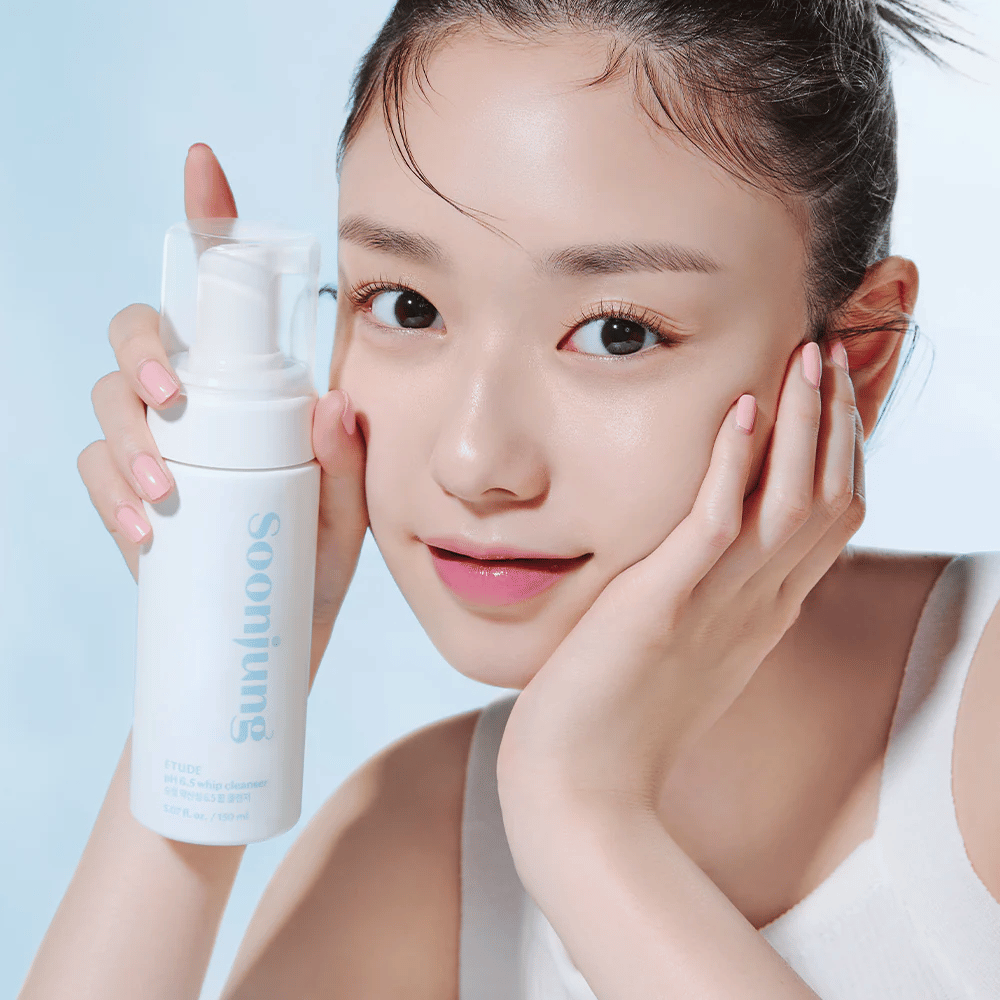Une jeune femme à la peau claire tient une bouteille pompe blanche avec le texte "Soon Jung pH 6.5 Whip Cleanser" de ETUDE. Elle sourit à la caméra avec un maquillage léger et une allure fraîche contre un fond bleu clair.