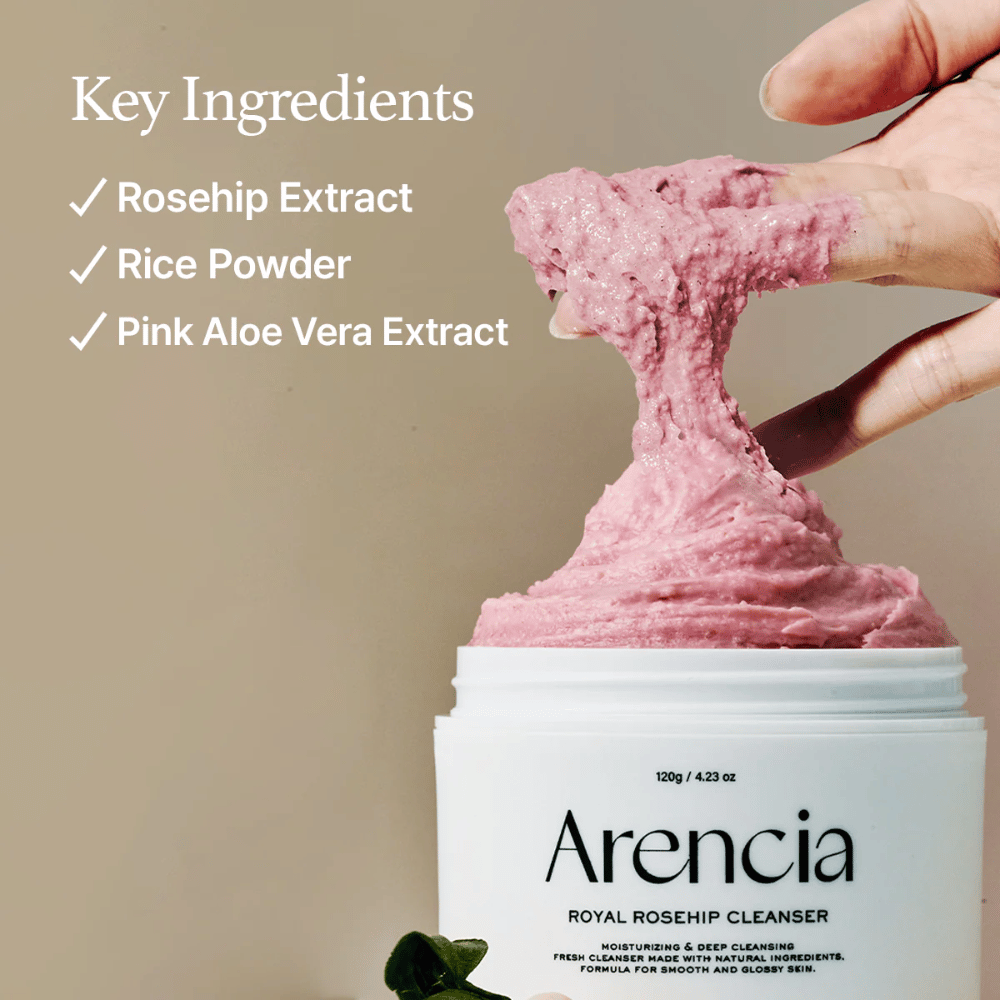 Une main soulève la texture nettoyante rose et moelleuse du pot Arencia Royal Rosehip Cleanser, avec la liste des ingrédients à gauche : extrait d'églantier, poudre de riz et extrait d'aloe vera rose.