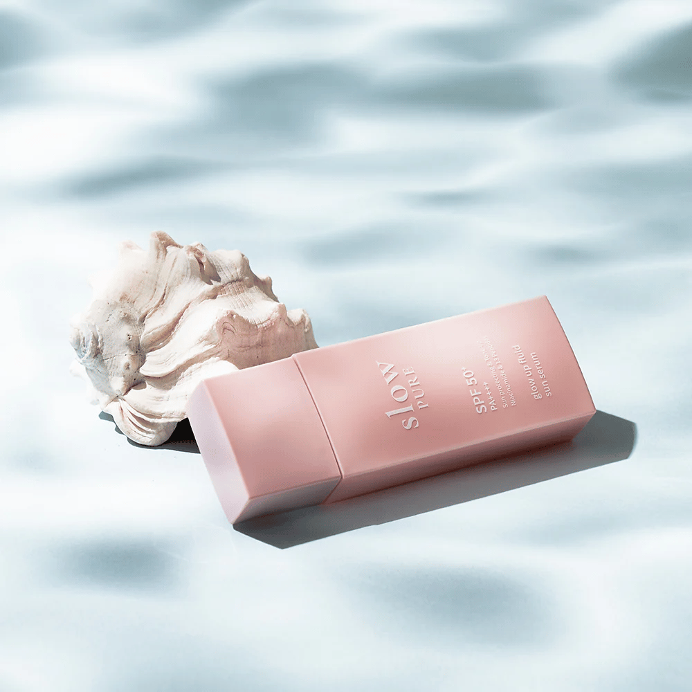 Slow Pure Glow Up Fluid Sun Serum près de coquillages et effet d'eau, met en avant la protection solaire et l'éclat.