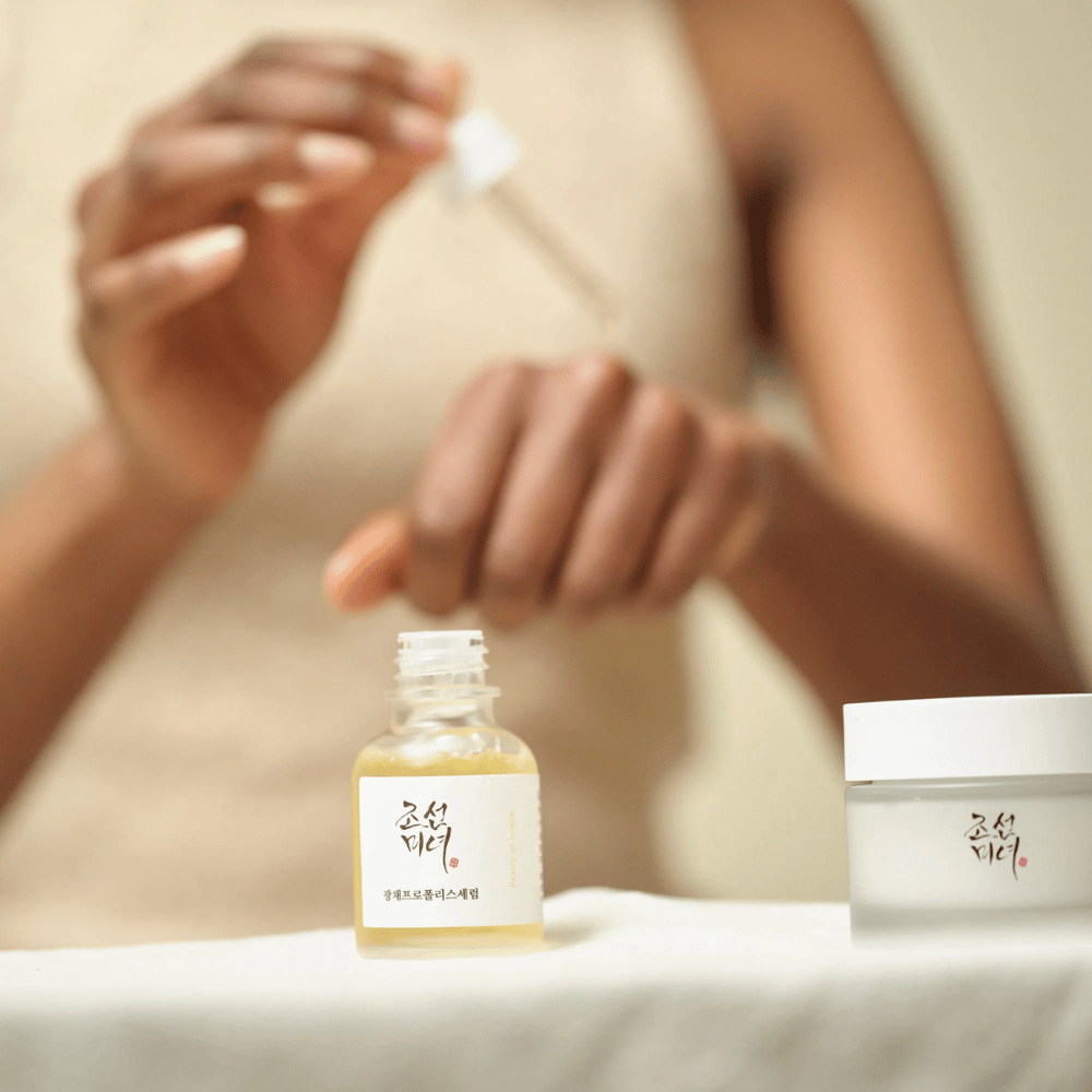 Personne appliquant Beauty of Joseon Glow Serum avec pipette à partir d'un flacon en verre, avec un pot de la même série à côté sur la table.